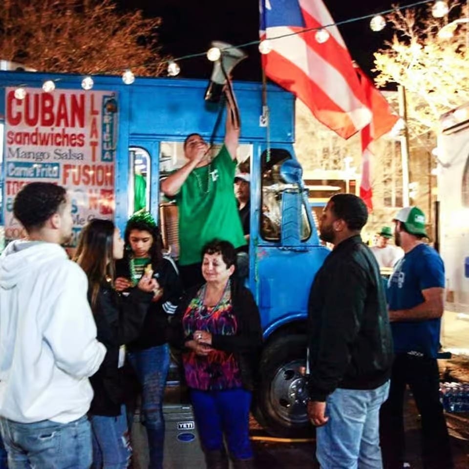 Crowd gathers around a bright blue Cuban sandwich food truck decorated with string lights and a large American flag.